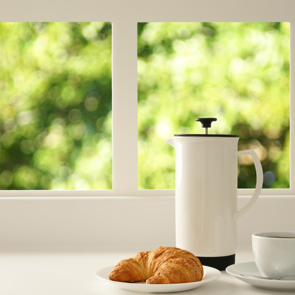 White French press coffee maker on a sunny balcony table with blurred greenery in the background
