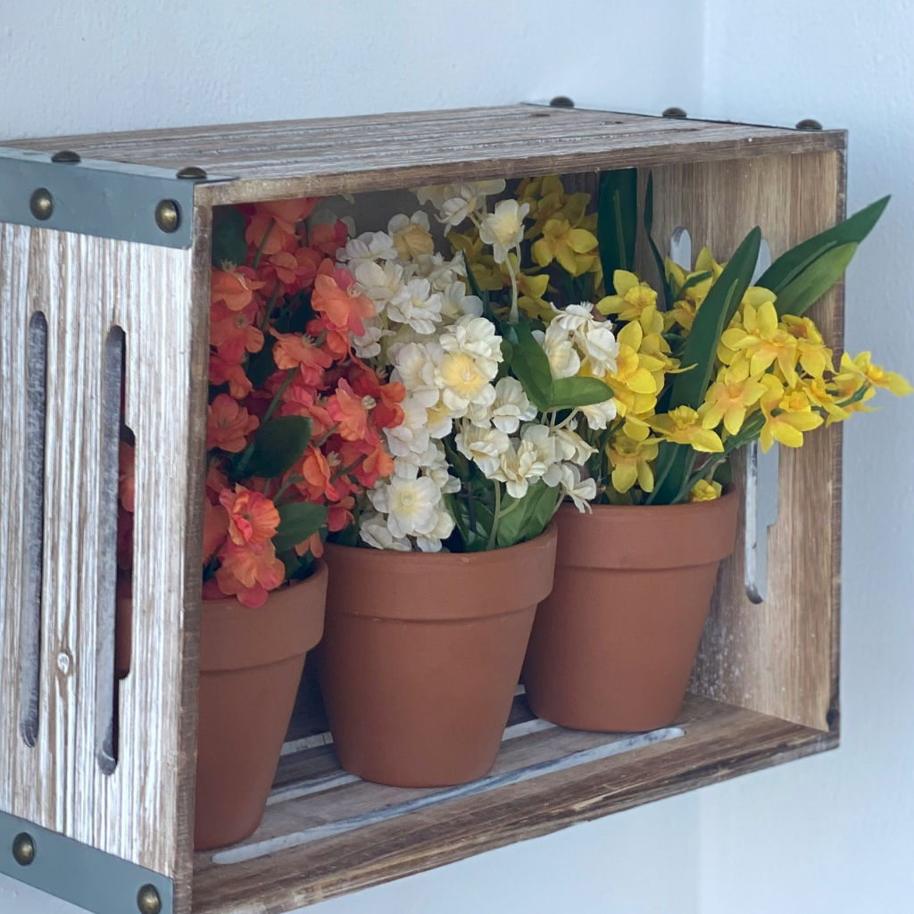 Colorful faux flowers in terracotta pots displayed in a rustic wooden crate on the wall