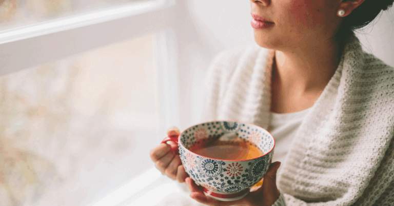 Woman holding a patterned ceramic bowl of tea near a window.