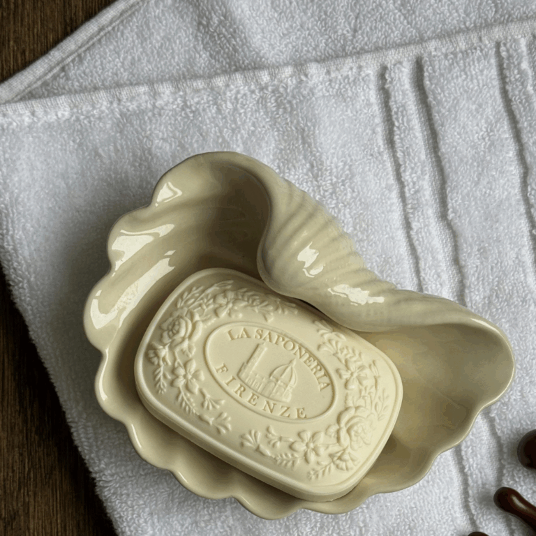 Beige seashell-shaped ceramic soap dish holding an embossed bar of soap on a white towel.