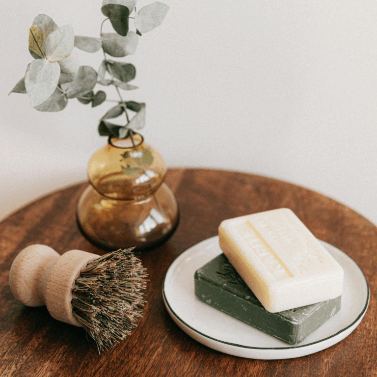 Two stacked natural soap bars, a wooden scrub brush, and an amber vase with eucalyptus on a wooden table.