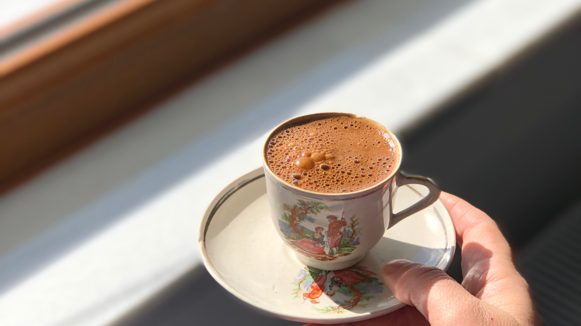 Hand holding a cup of coffee on a colorful patterned saucer, styled with cozy blanket and dried flowers, a calming morning ritual idea from the blog "3 Mini Rituals You Can Create in Under 5 Minutes (Using Everyday Decor).