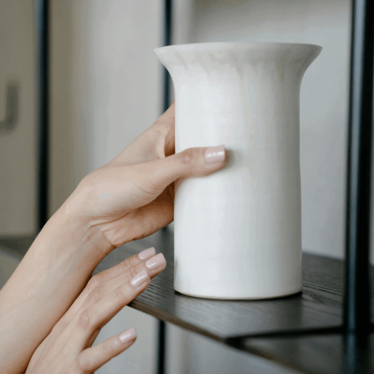 Hands carefully placing a minimalist white ceramic vase on a dark wooden shelf for home decor