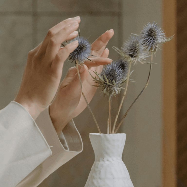 Hands arranging dried thistle flowers in a textured white ceramic vase for interior styling