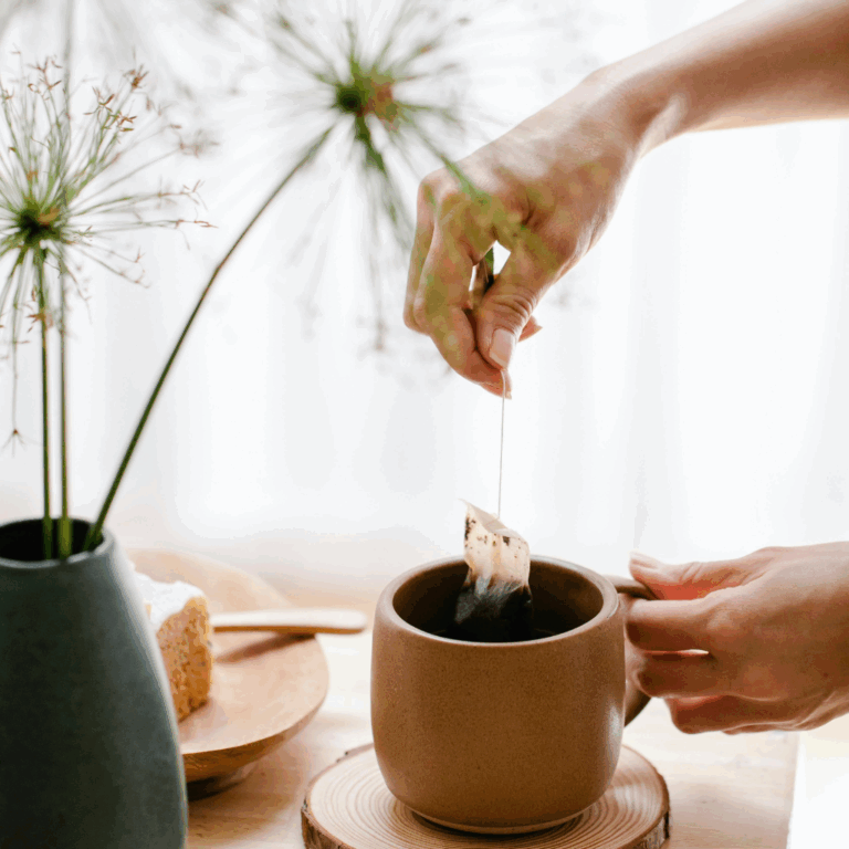 Hand dipping a tea bag into a brown ceramic mug beside decorative plants and wooden tray