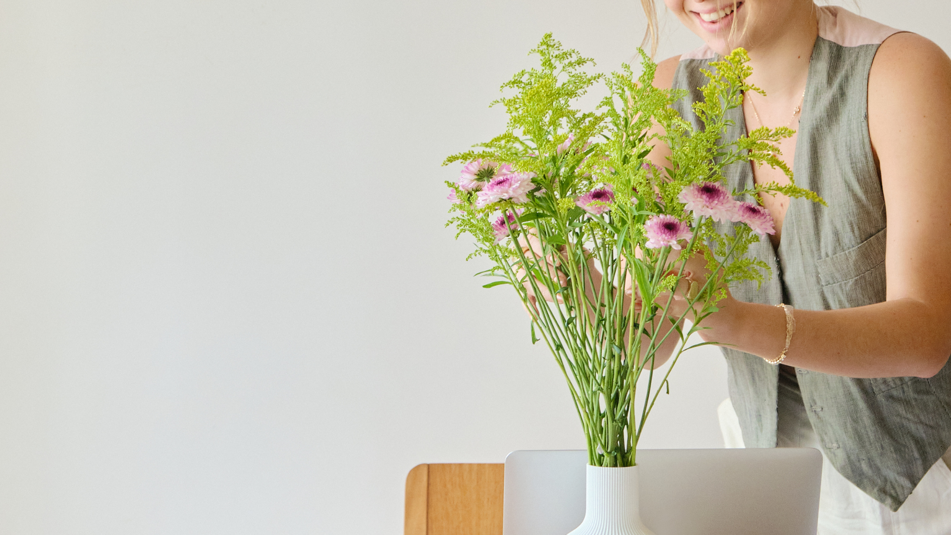 Smiling woman arranging pink flowers in a white vase on a work desk