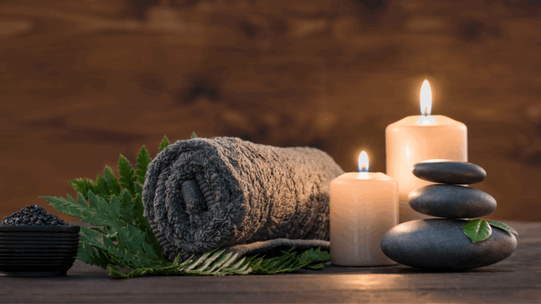Rolled gray towel, burning candles, black massage stones, and fresh green fern leaves arranged on a wooden surface for a calming spa atmosphere.