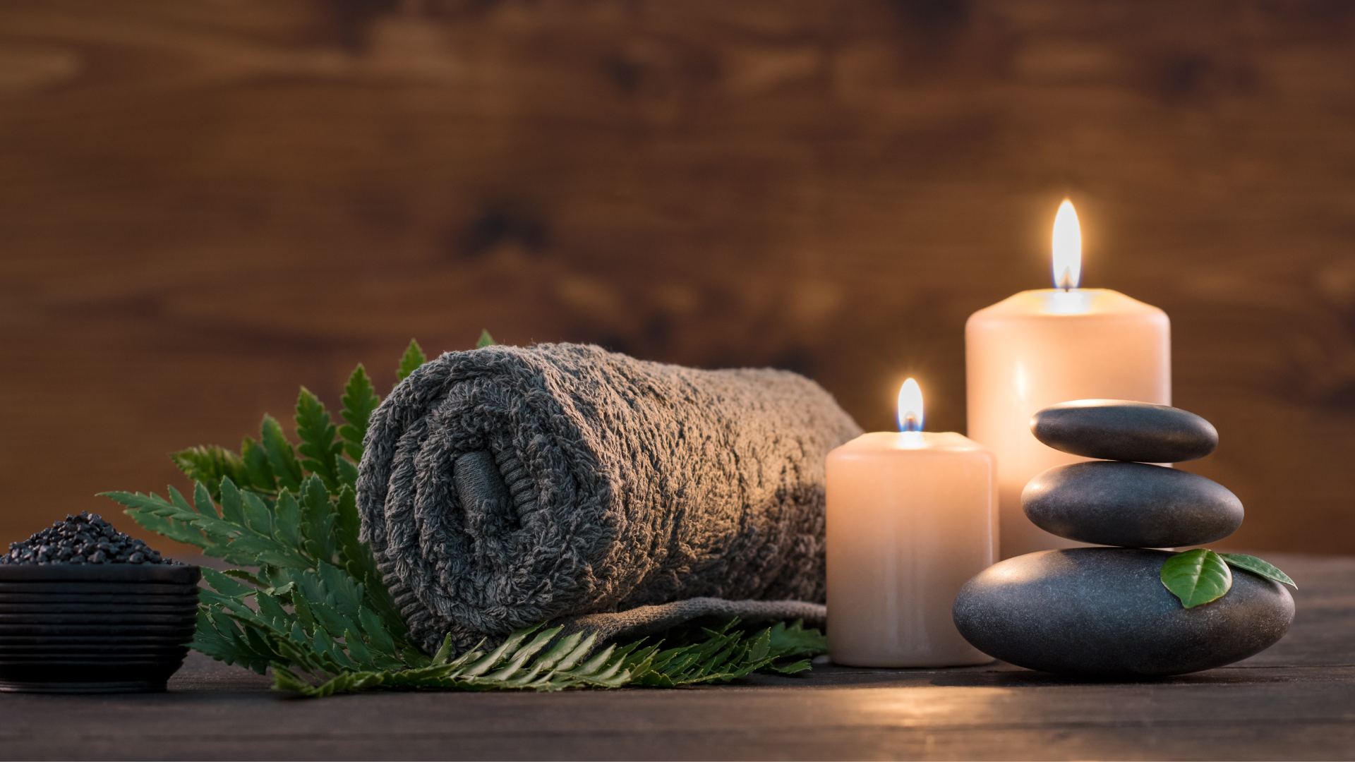 Rolled gray towel, burning candles, black massage stones, and fresh green fern leaves arranged on a wooden surface for a calming spa atmosphere.