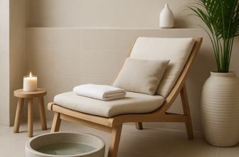 Minimalist spa corner with a beige lounge chair, soft cushion, rolled towel, wooden stool with a lit candle, potted palm plant, and a round stone foot basin, styled for a calming home retreat.