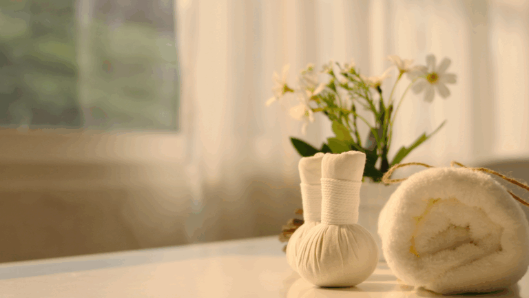 Rolled white towel, herbal massage compress balls, and chamomile flowers placed on a clean surface with soft natural light—ideal for a calming spa nook.