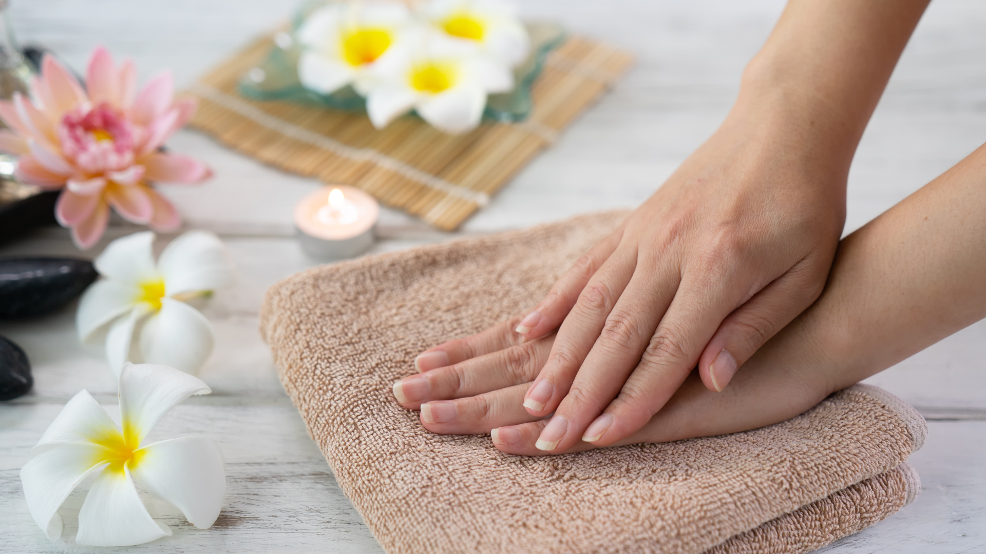 Hands gently massaging on a soft brown towel surrounded by white plumeria flowers, pink lotus, black stones, a tealight candle, and spa decor on a white wooden table.