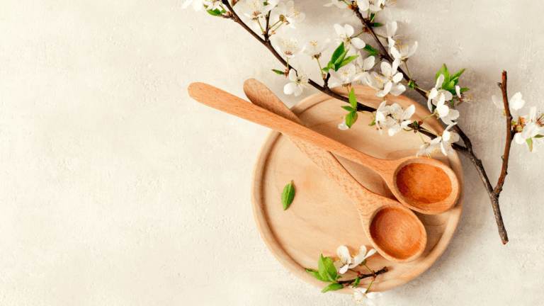 Two wooden spoons on a round wooden plate, surrounded by blooming white cherry blossoms on a neutral textured background—ideal for natural spa or wellness themes.