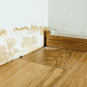 Water damage on a lower interior wall with paint peeling above a wooden floor.