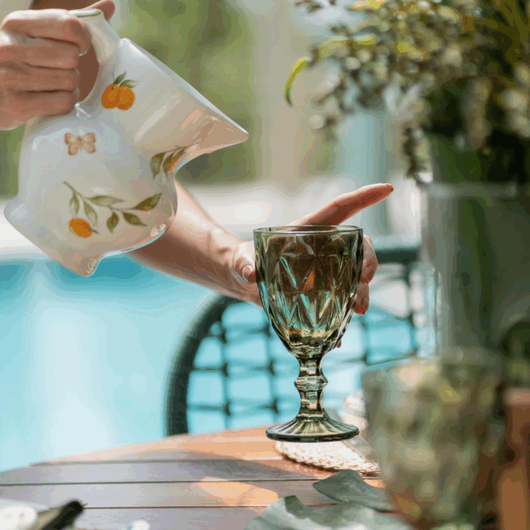 Hand pouring a drink from a white ceramic pitcher with citrus design into a green glass