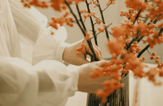 Hands arranging orange berry branches in a tall dark vase on a wooden table.