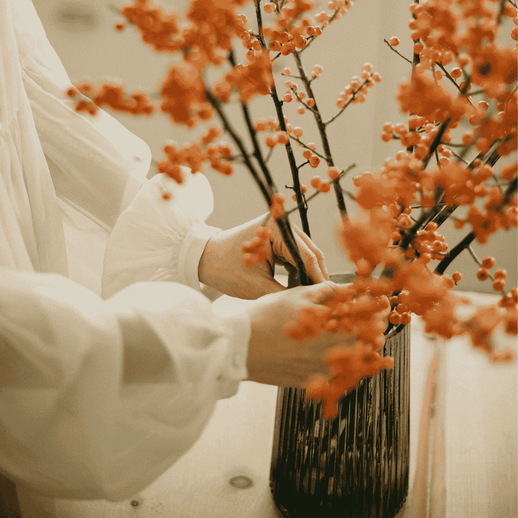 Hands arranging orange berry branches in a tall dark vase on a wooden table.