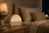 Hatch Restore 3 sunrise alarm clock glowing on a wood nightstand at night beside a linen bed and warm lamp.