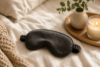 Black silk sleep mask on linen bedding with eucalyptus, candle, and wood tray in a warm minimalist bedroom.