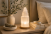 Tiered selenite tower lamp glowing softly on a wooden base, styled on a nightstand with a ceramic vase, dried sage, and stacked books